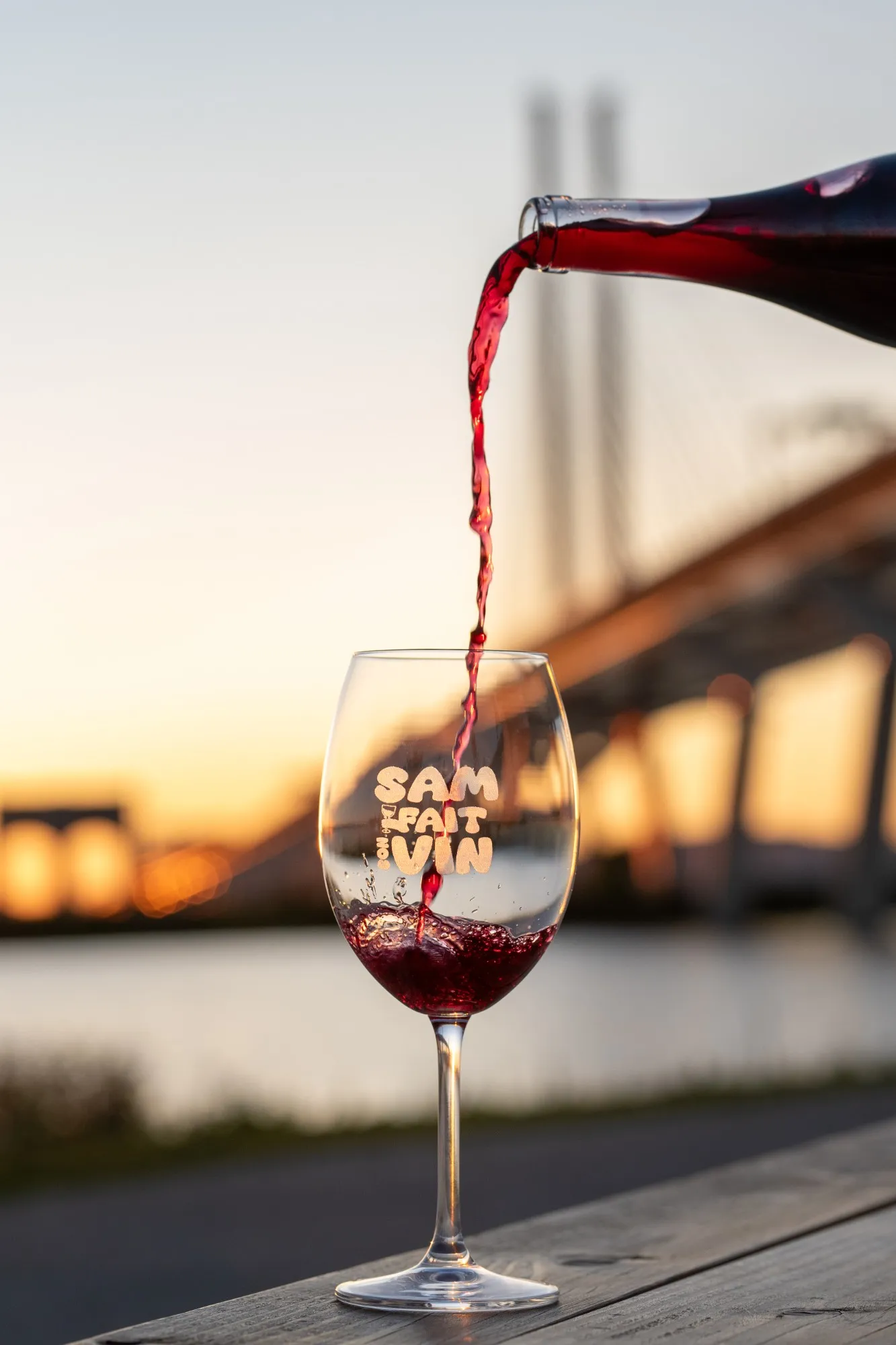 Du vin rouge versé d’une bouteille dans un verre gravé du logo Sam fait son vin, au coucher du soleil, devant un pont flou